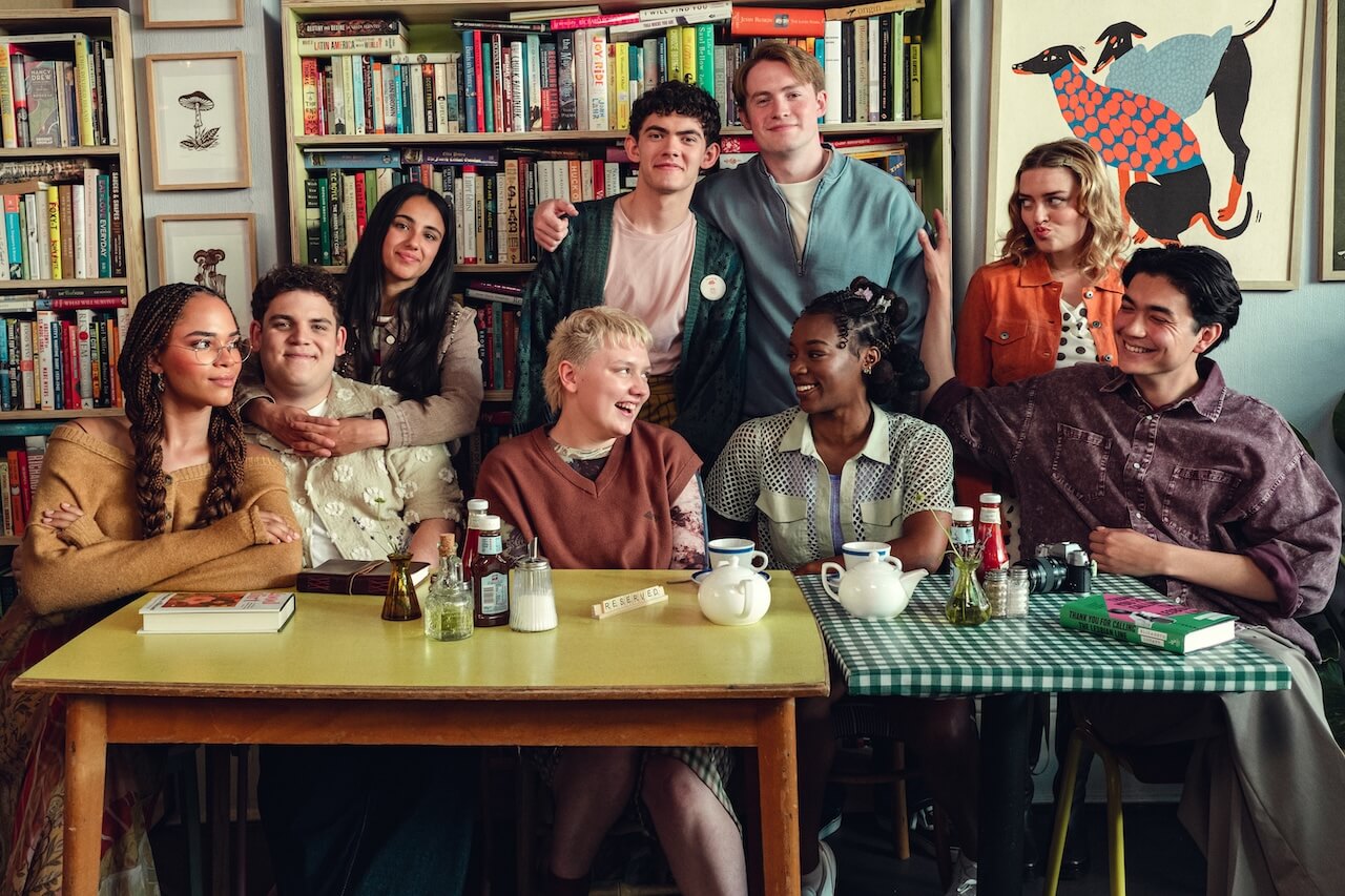 "Nine young adults sit and stand around a table in a cozy cafe or library, surrounded by bookshelves and artwork, enjoying tea and conversation, with condiments, teapots, and cups visible on the tables."