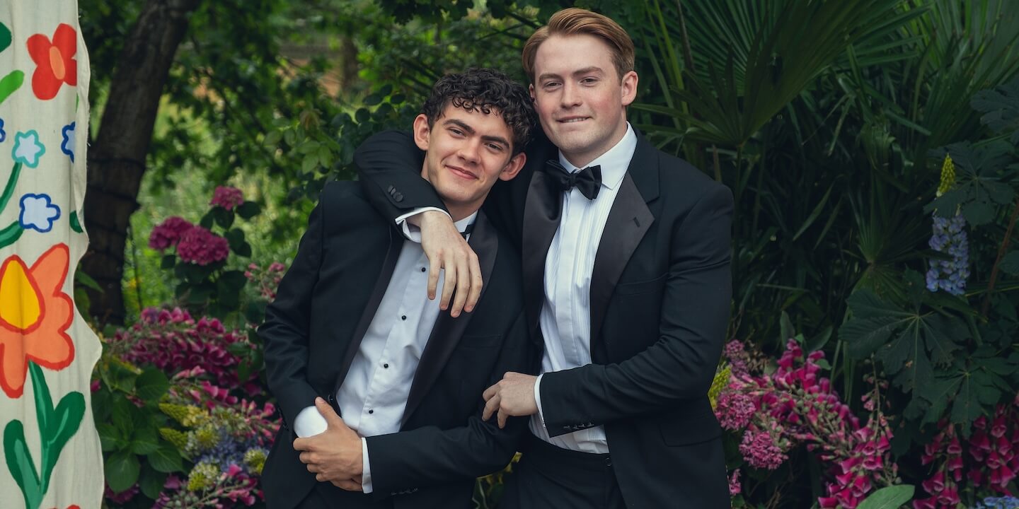 "Two young men in black tuxedos pose together outdoors, standing close with one arm around the other, smiling in a lush garden surrounded by vibrant flowers and greenery, with a colorful floral backdrop to the left."